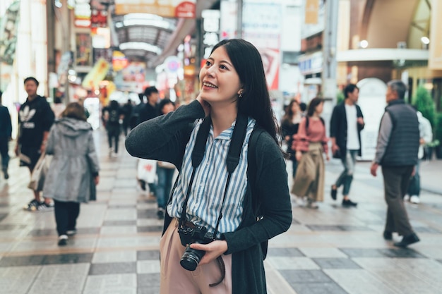 hermosa joven sosteniendo la cámara de pie en la calle en la famosa zona comercial de osaka, japón. viajero visitando el puente shinsaibashi sonriendo relajándose en la carretera bajo techo. multitud de gente local caminando