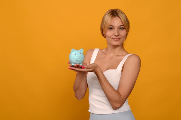 Hermosa joven sosteniendo una hucha de cerdo sobre fondo amarillo. Para ahorrar dinero, riqueza y concepto financiero.