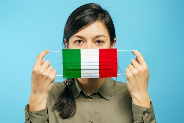 Hermosa joven sosteniendo una máscara protectora médica en sus manos con la bandera nacional italiana sobre un fondo azul.