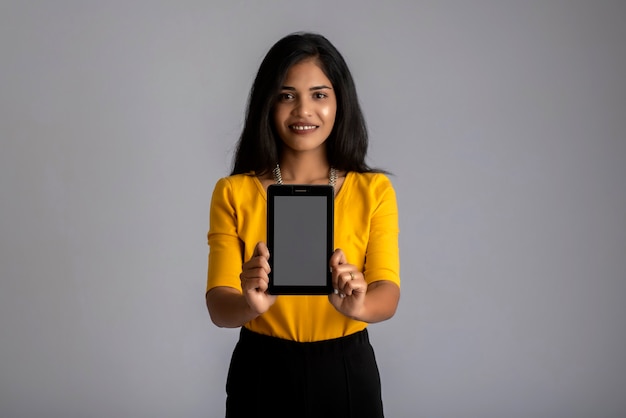 Hermosa joven sosteniendo y mostrando la pantalla en blanco del teléfono inteligente o teléfono móvil o tableta en una pared gris.