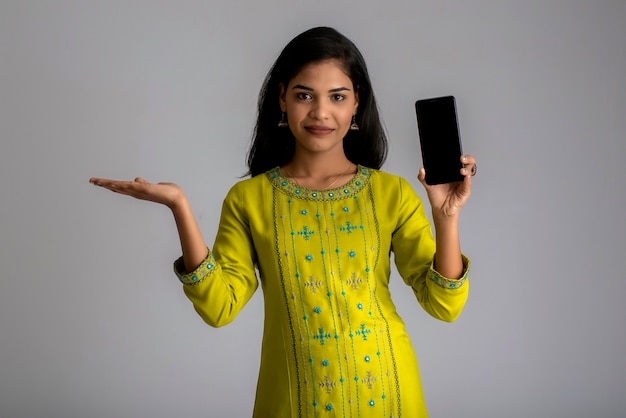 Hermosa joven sosteniendo y mostrando la pantalla en blanco del teléfono inteligente o teléfono móvil o tableta en una pared gris.