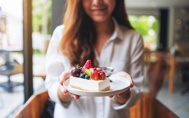 Una hermosa joven sosteniendo y mostrando un plato de tarta de queso de frutas mixtas en el café