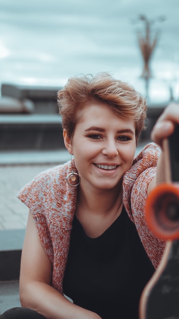 Una hermosa joven sosteniendo una patineta mientras está sentada en el skatepark en la ciudad