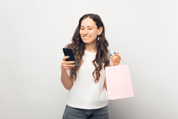 Foto hermosa joven sostiene una bolsa de compras, mientras ordena algo o paga por teléfono.