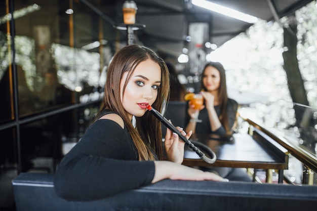 Hermosa joven con su novia, se sienta y fuma una cachimba en la terraza de verano de un café moderno.