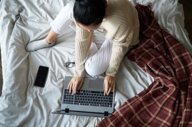 Hermosa joven en suéter acogedor en la cama en casa trabajando en una computadora portátil