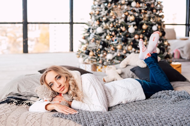 Una hermosa joven con un suéter blanco se encuentra en el sofá cerca de un hermoso árbol de Navidad