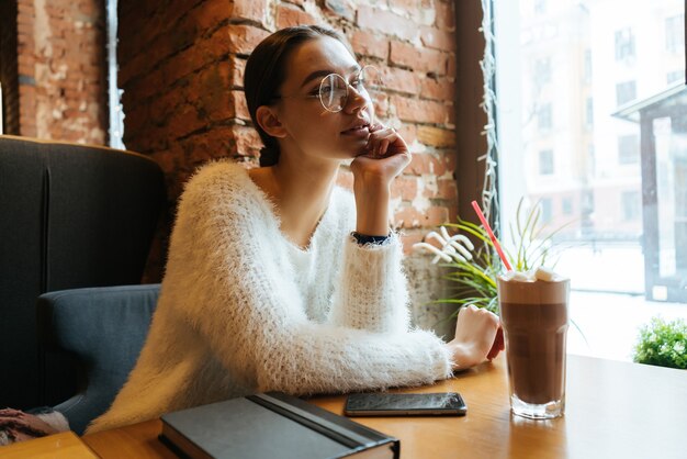 Hermosa joven con un suéter blanco y gafas se sienta en un café y mira pensativamente por la ventana