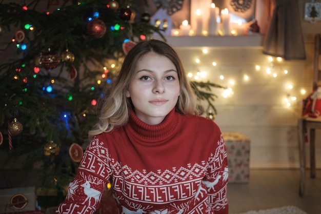 Hermosa joven de suéter rojo en el árbol de Navidad. Interior festivo, velas, luces, guirnaldas. Retrato de una niña en la noche de Navidad