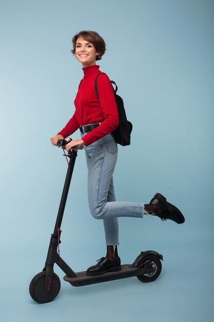 Foto hermosa joven en suéter rojo y jeans con mochila negra de pie con scooter eléctrico mientras mira alegremente en cámara sobre fondo azul.