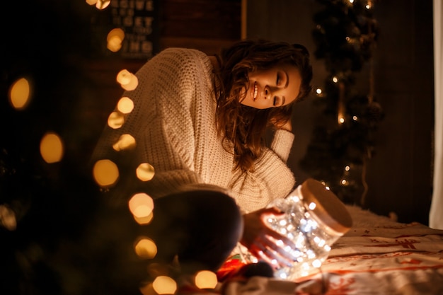 Hermosa joven en un suéter vintage de punto con un frasco mágico con luces festivas en una cama en Nochebuena