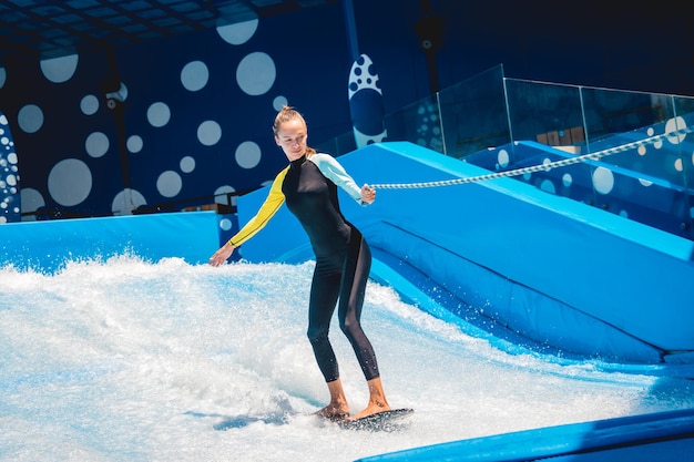 Hermosa joven surfeando con un entrenador en un simulador de olas en un parque de atracciones acuático