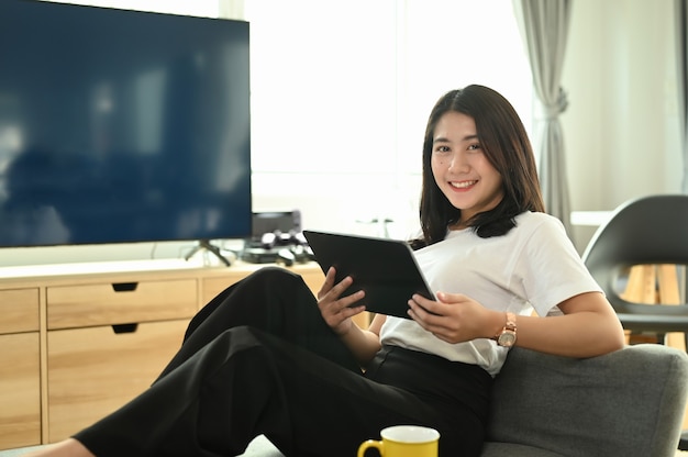 Hermosa joven con tableta digital y sonriendo mientras está sentado en el sofá en casa.