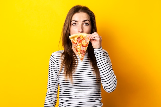 Hermosa joven tapándose la boca con una rebanada de pizza fresca en un amarillo