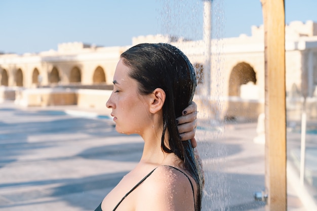 Hermosa joven toma una relajante ducha en traje de baño en un día soleado al aire libre junto al mar.