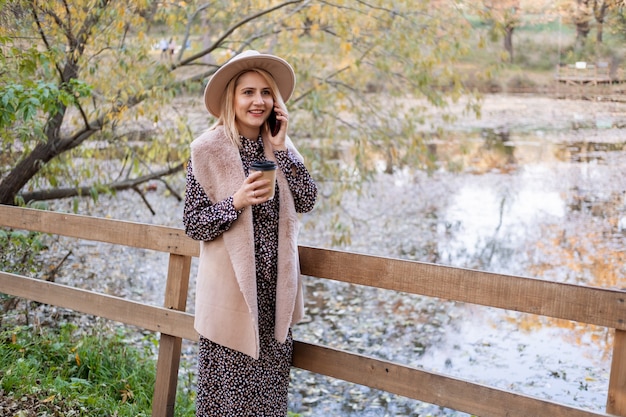 Hermosa joven tomando café, hablando por teléfono en la naturaleza junto al lago en el parque de otoño en otoño.