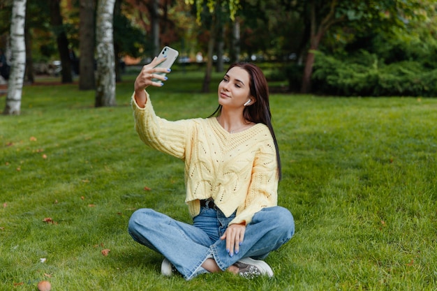 Hermosa joven tomando una foto selfie en el parque con un teléfono inteligente. chica con un teléfono sentada en el césped del parque.