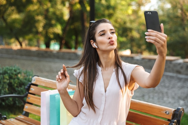 Hermosa joven tomando un selfie mientras está sentado en un banco con bolsas de compras al aire libre