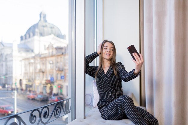 Hermosa joven tomando un selfie con teléfono en el alféizar de la ventana del hotel frente a la ciudad