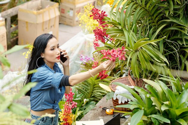 Hermosa joven trabajadora de vivero de flores asiáticas hablando por teléfono con el cliente que está pidiendo arbusto con flores rojas para el patio trasero