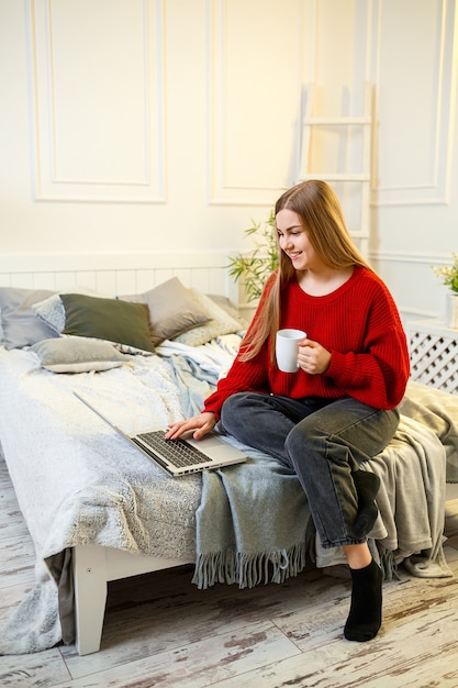 Hermosa joven trabajando en una computadora portátil sentada en la cama en su casa, ella está tomando café y sonriendo. Trabaja desde casa durante la cuarentena. Chica en un suéter y jeans en casa en la cama