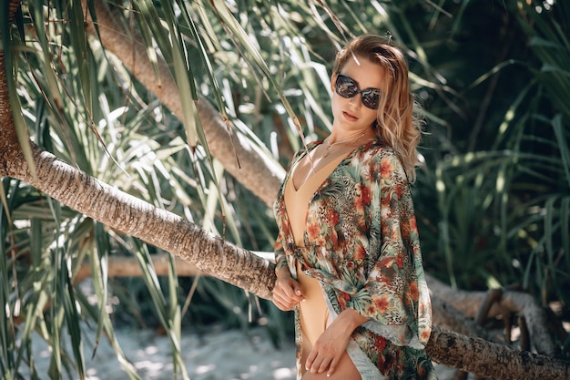 Hermosa joven en traje de baño beige y capa de flores y gafas de sol negras posando en la playa cerca de la palmera