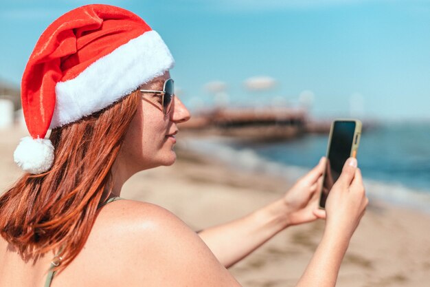 Hermosa joven en traje de baño y sombrero rojo de santa claus hace un selfie en la orilla del mar