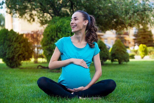 Hermosa joven tranquila embarazada está sentada en la posición de loto y meditando mientras practica yoga en el parque