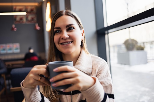 Hermosa joven tranquila y sonriente bebiendo café mirando por la ventana mientras se sienta en una mesa en un restaurante. Cafe mañanero