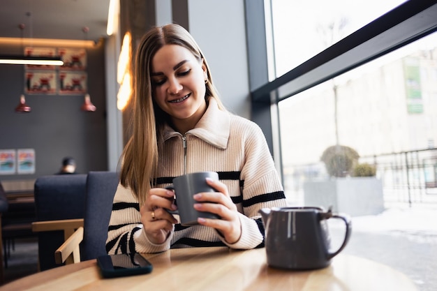 Hermosa joven tranquila y sonriente bebiendo café mirando por la ventana mientras se sienta en una mesa en un restaurante. Cafe mañanero