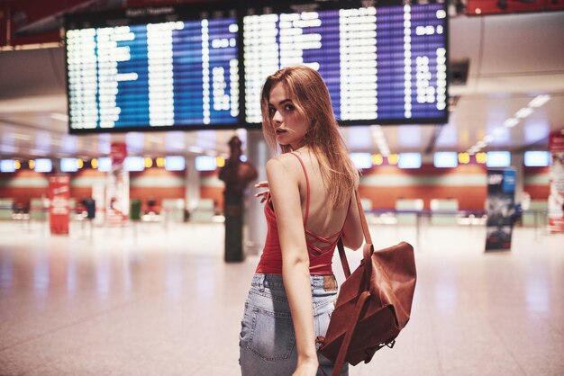 Hermosa joven turista con mochila en el aeropuerto internacional, cerca del tablero de información de vuelo.