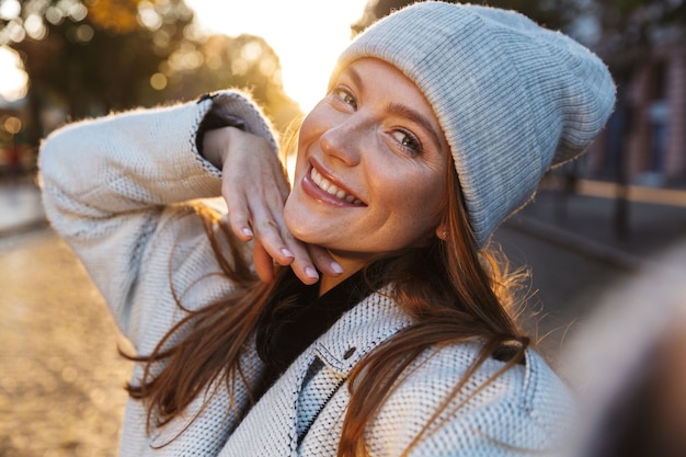 Hermosa joven vestida con abrigo y sombrero de otoño caminando al aire libre, tomando un selfie