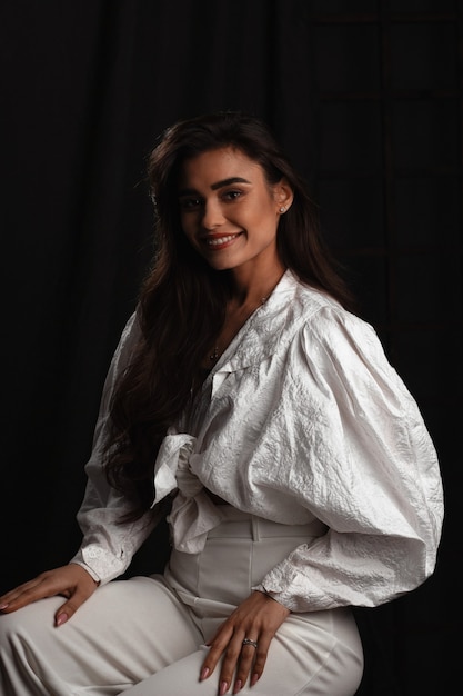 Hermosa joven vestida de blanco posando en un estudio fotográfico oscuro. Foto de bajo perfil. Sensible mujer de cabello castaño con cabello largo. Mujer joven con hermosa sonrisa.