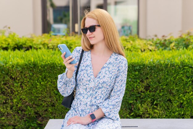 Una hermosa joven vestida está sentada en un banco y escribiendo en su teléfono