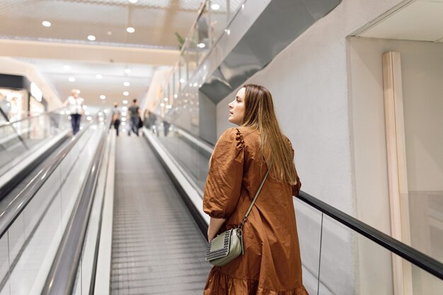 Hermosa joven vestida de material natural color chocolate con leche se eleva en la escalera mecánica y mira a su alrededor. Compras y entretenimiento. Recreación y viajes. Vida cómoda.