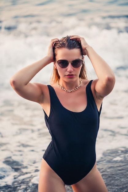 Hermosa joven vestida con un traje de baño negro se arrodilla en la playa de arena Enfoque selectivo