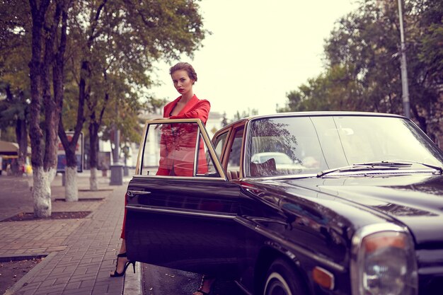 Hermosa joven vestida con traje rojo mientras está de pie cerca de coche retro