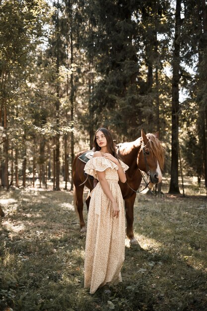 Una hermosa joven vestida con un vestido se encuentra cerca de un caballo en el bosque