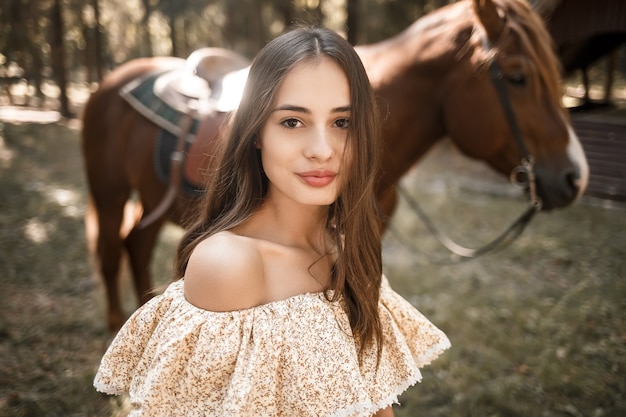 Una hermosa joven vestida con un vestido se encuentra cerca de un caballo en el bosque