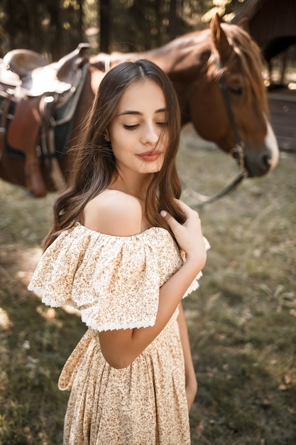 Una hermosa joven vestida con un vestido se encuentra cerca de un caballo en el bosque