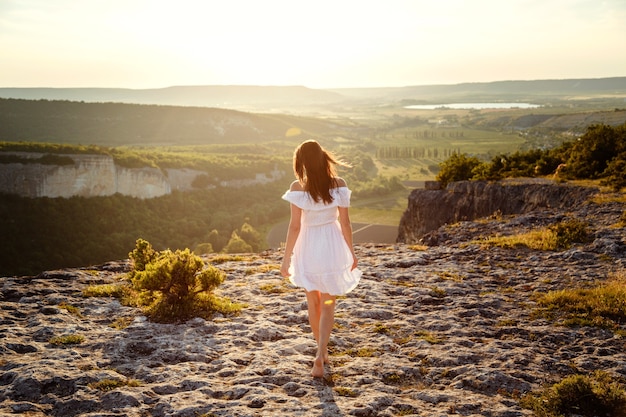Hermosa joven con un vestido blanco disfruta de un paisaje impresionante en las montañas durante la puesta de sol