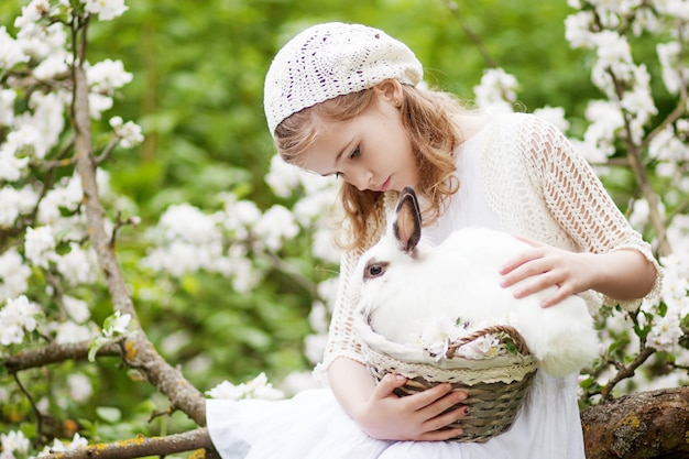 Hermosa joven con un vestido blanco jugando con conejo blanco en el jardín de flores de primavera. Actividad divertida de primavera para niños. tiempo de Pascua