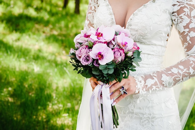 Hermosa joven en vestido elegante está de pie y sosteniendo la mano ramo de flores de color rosa pastel y verdes con cinta en la naturaleza La novia sostiene un ramo de novia al aire libre