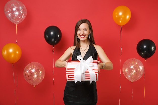 Hermosa joven en vestido negro dando o recibiendo, sosteniendo una caja roja con regalo, presente en globos de aire de fondo rojo brillante. Día de la mujer, feliz año nuevo, concepto de fiesta de vacaciones de maqueta de cumpleaños.