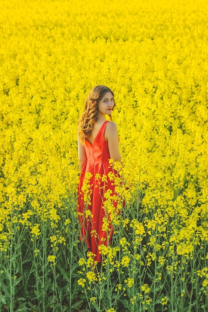 Hermosa joven con un vestido rojo de cerca en medio del campo amarillo con el primer plano de flores de rábano.