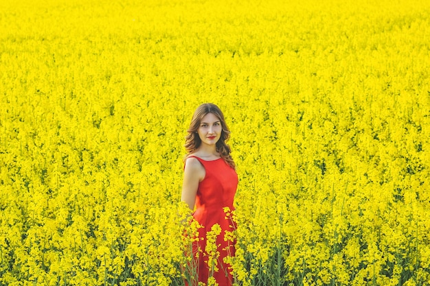 Hermosa joven con un vestido rojo de cerca en medio del campo amarillo con el primer plano de flores de rábano.