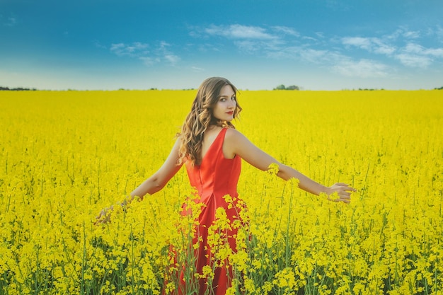 Hermosa joven con un vestido rojo de cerca en medio del campo amarillo con el primer plano de flores de rábano.