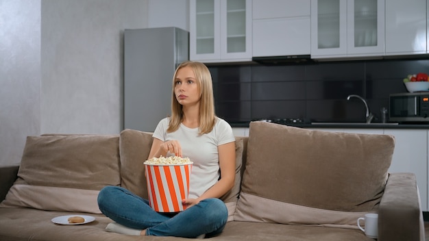Hermosa joven viendo una película y sentada en el sofá con palomitas de maíz.