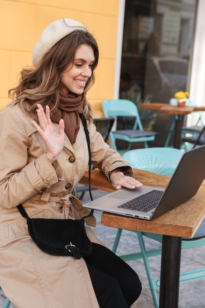 Hermosa joven vistiendo un abrigo usando una computadora portátil mientras está sentado en el café al aire libre, agitando la mano