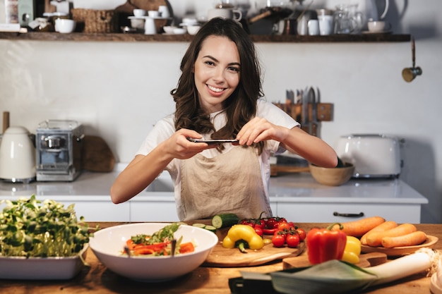 Hermosa joven vistiendo delantal cocinar ensalada saludable en la cocina de casa, tomando una foto de la comida con el teléfono móvil